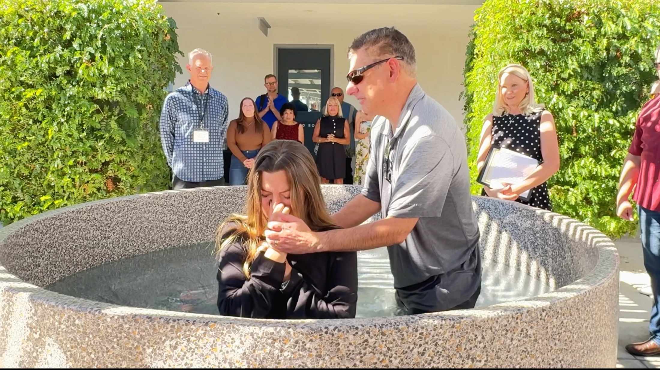 A pastor baptizes a woman in a circular stone font as a small crowd watches. The ceremony is taking place outdoors on a sunny day.